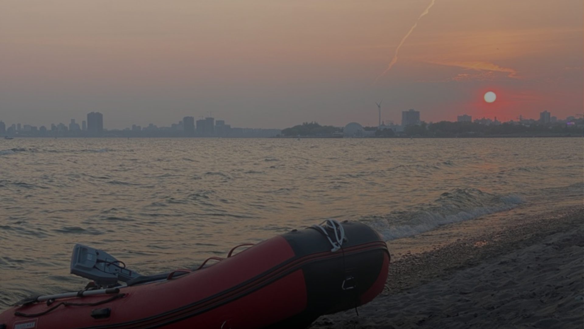 A red inflatable boat sits on a beach at sunset, a hazy city skyline visible across the water.