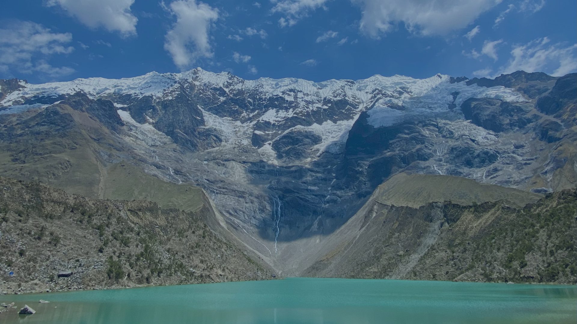A turquoise glacial lake sits beneath a wall of snow-covered mountain peaks.