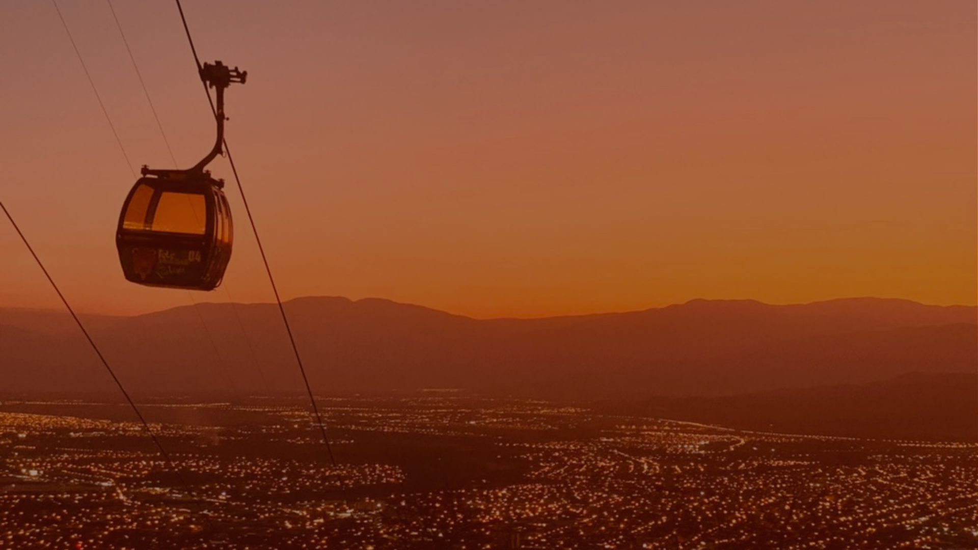 A cable car descends a line toward a city lit up at dusk, mountains rising behind it.