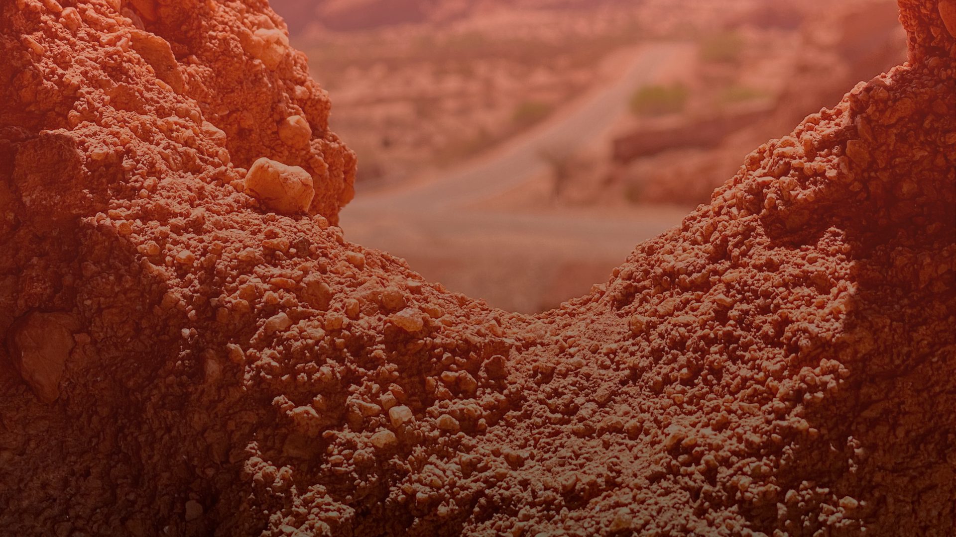 Close view of rough red-orange earth and small stones forming a curved gap, through which a blurred desert road winds off into the distance.