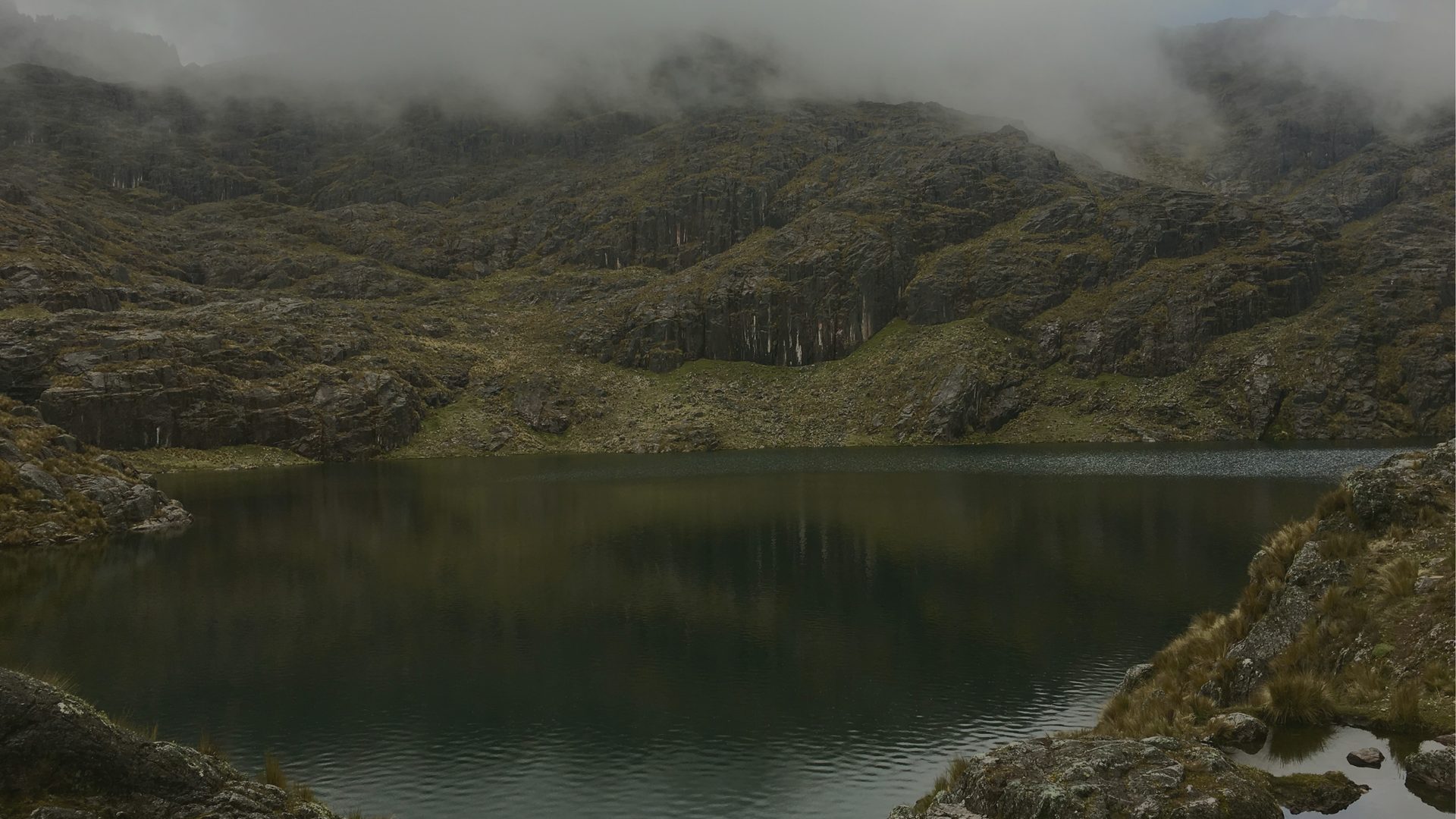 A still mountain lake mirrors steep, forested cliffs wrapped in low cloud.