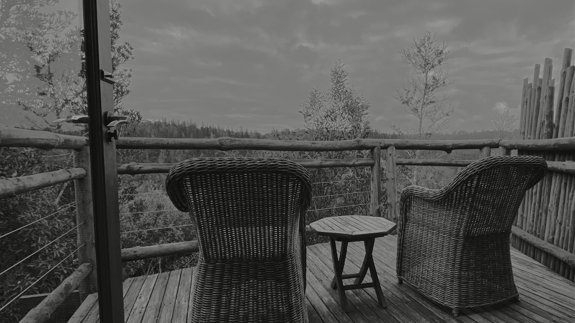 Two empty wicker chairs and a small table sit on a wooden deck overlooking a forest.