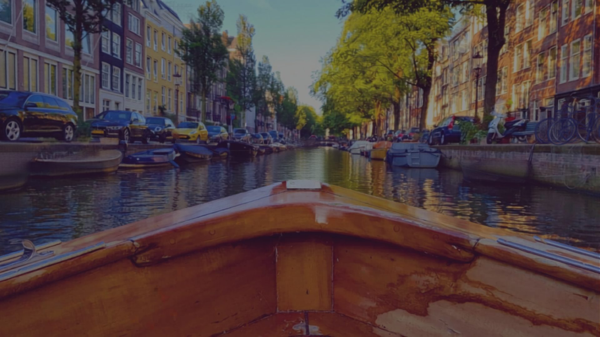 The varnished bow of a wooden boat leads toward a canal lined with parked cars, houseboats, and old townhouses.