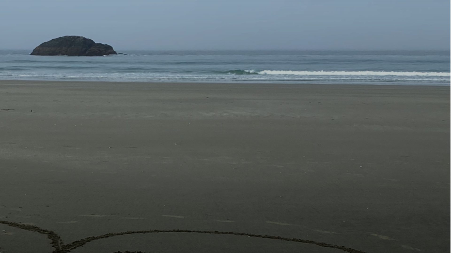 A grey sand beach under a hazy blue-grey sky, with a dark rock island offshore on the left, a line of surf along the shore, and a curved line drawn in the sand in the foreground.