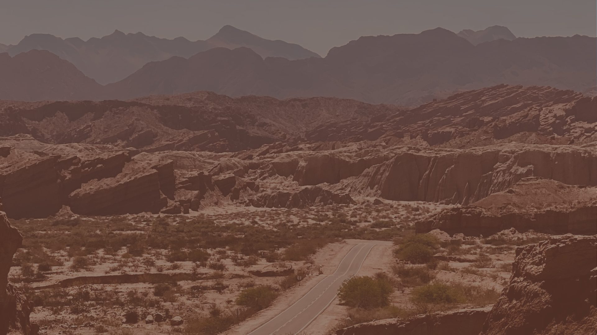 A dirt road curves through a canyon of layered red and grey rock under a hazy sky.