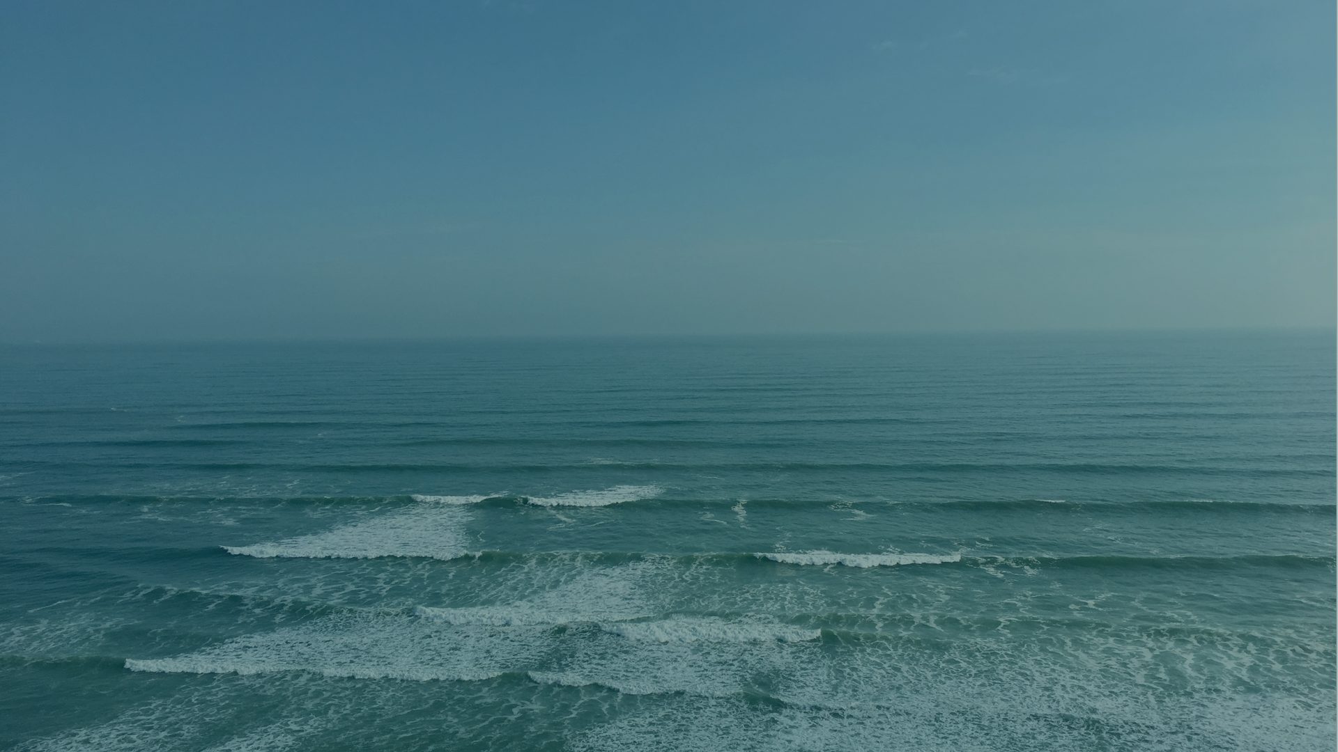 A wide stretch of ocean rolls in small waves toward the shore under a pale, even sky.