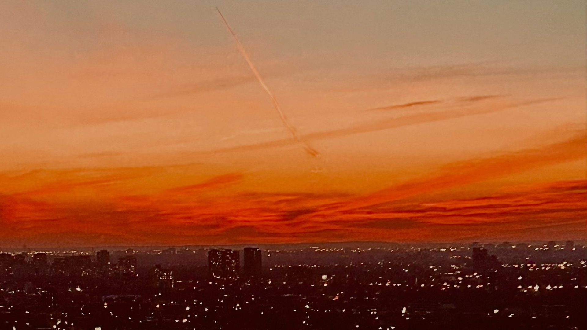 A jet's contrail crosses a fiery orange sunset sky above a city skyline.