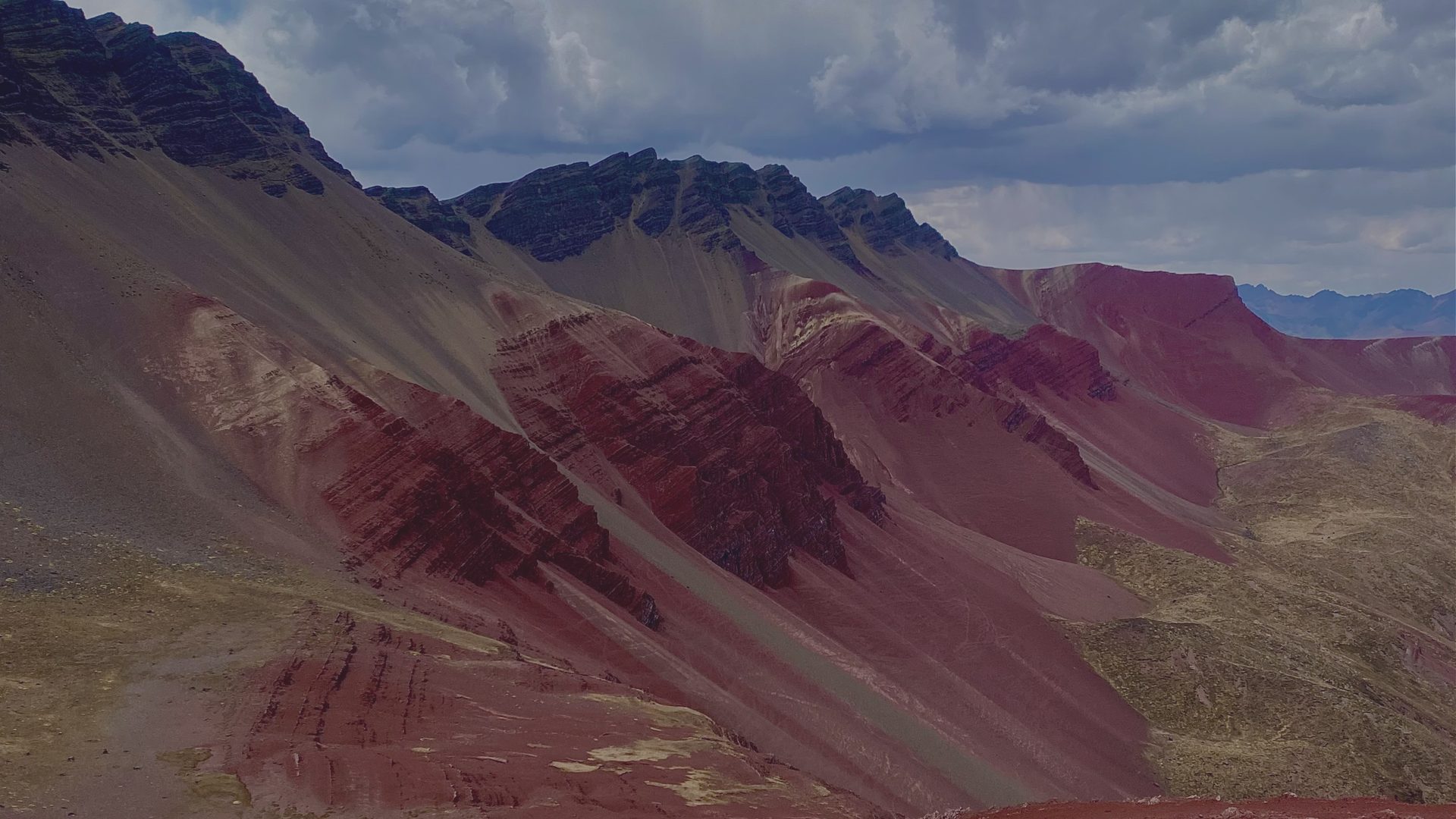 Striped red and maroon mountain slopes rise under a heavy, cloud-covered sky.
