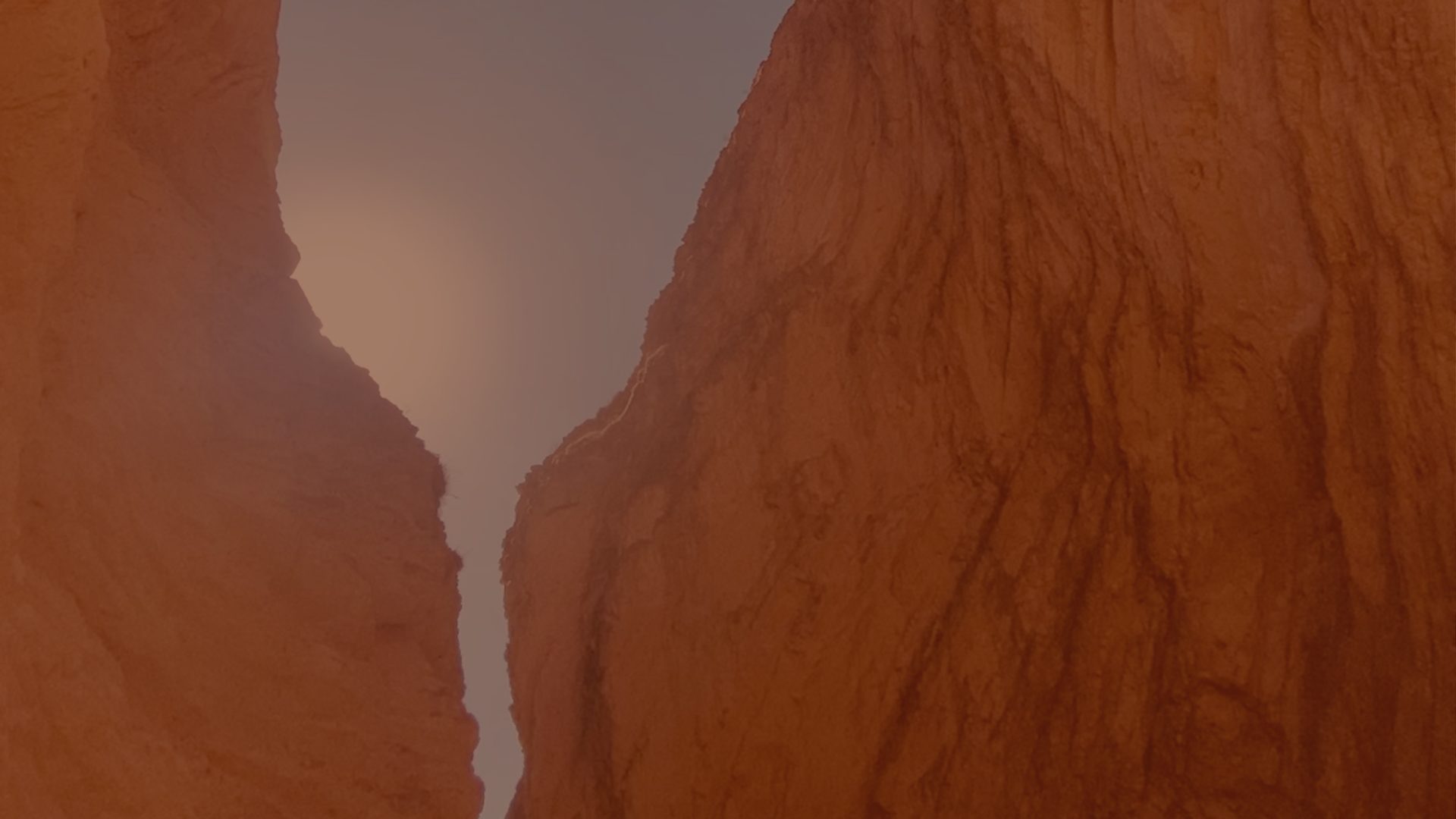 Looking up through a narrow gap between two rounded orange rock walls toward a pale, hazy sky.
