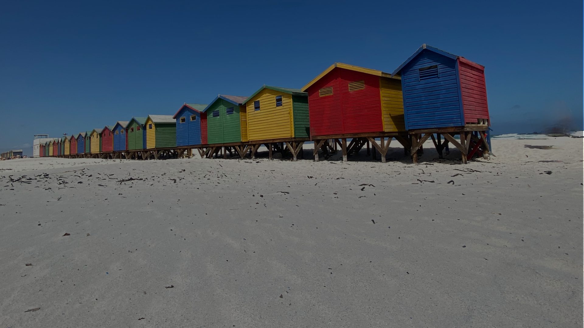 A row of brightly painted wooden beach huts on stilts, in blue, red, yellow and green, receding along a pale sandy beach under a deep blue sky.