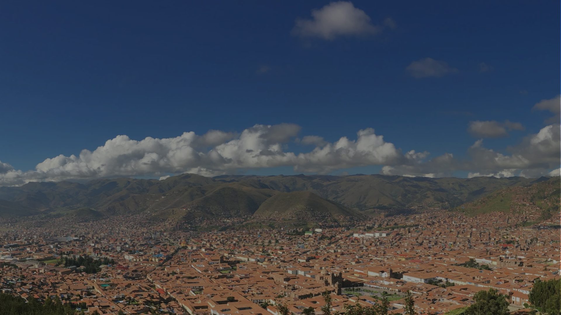 A sprawling terracotta-roofed city fills a Andean valley beneath green mountains, with a bank of white cumulus cloud against a deep blue sky.