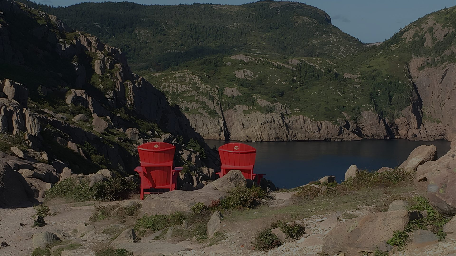 Two red chairs sit on a rocky cliff edge overlooking a narrow, dark fjord.