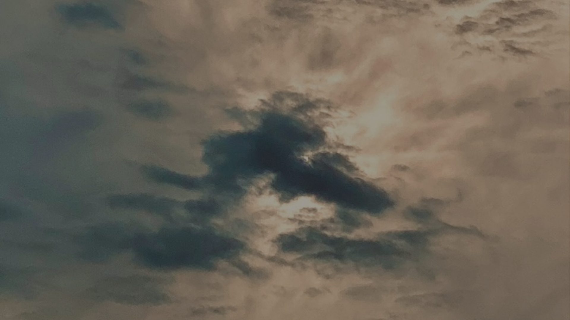 A dark cloud breaks apart against a hazy, sunlit sky.