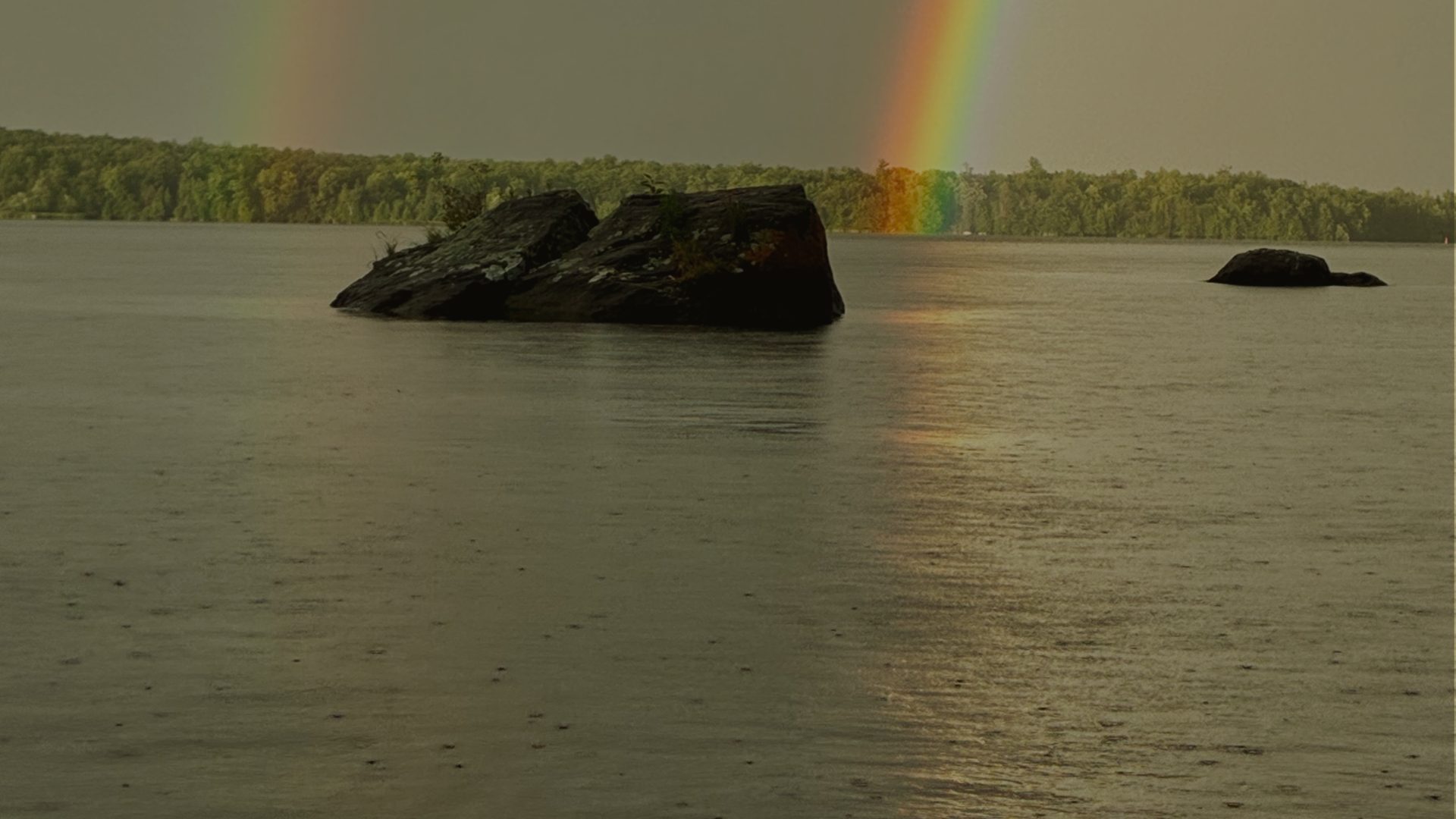 A dark rock outcrop rises from a still, rain-flecked lake under a grey sky, with a rainbow touching the tree line behind it.