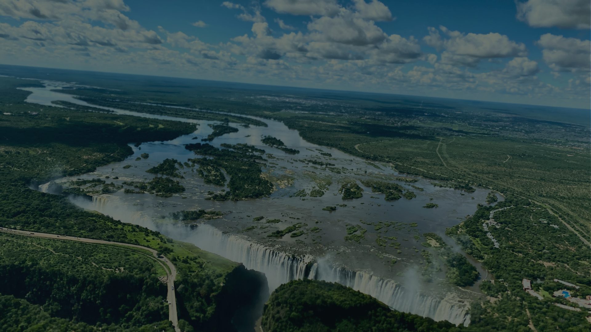 An aerial view of a wide waterfall spilling across a river gorge, mist rising over the flat green land beyond.