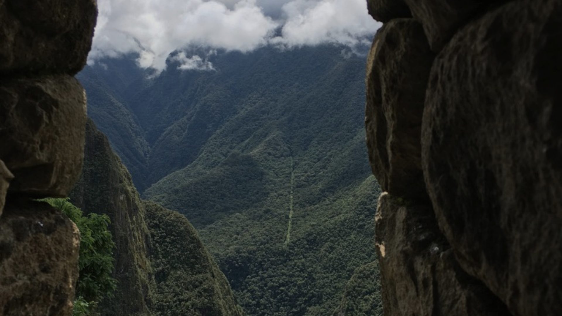 A gap between two rock faces opens onto a forested mountain gorge under low cloud.