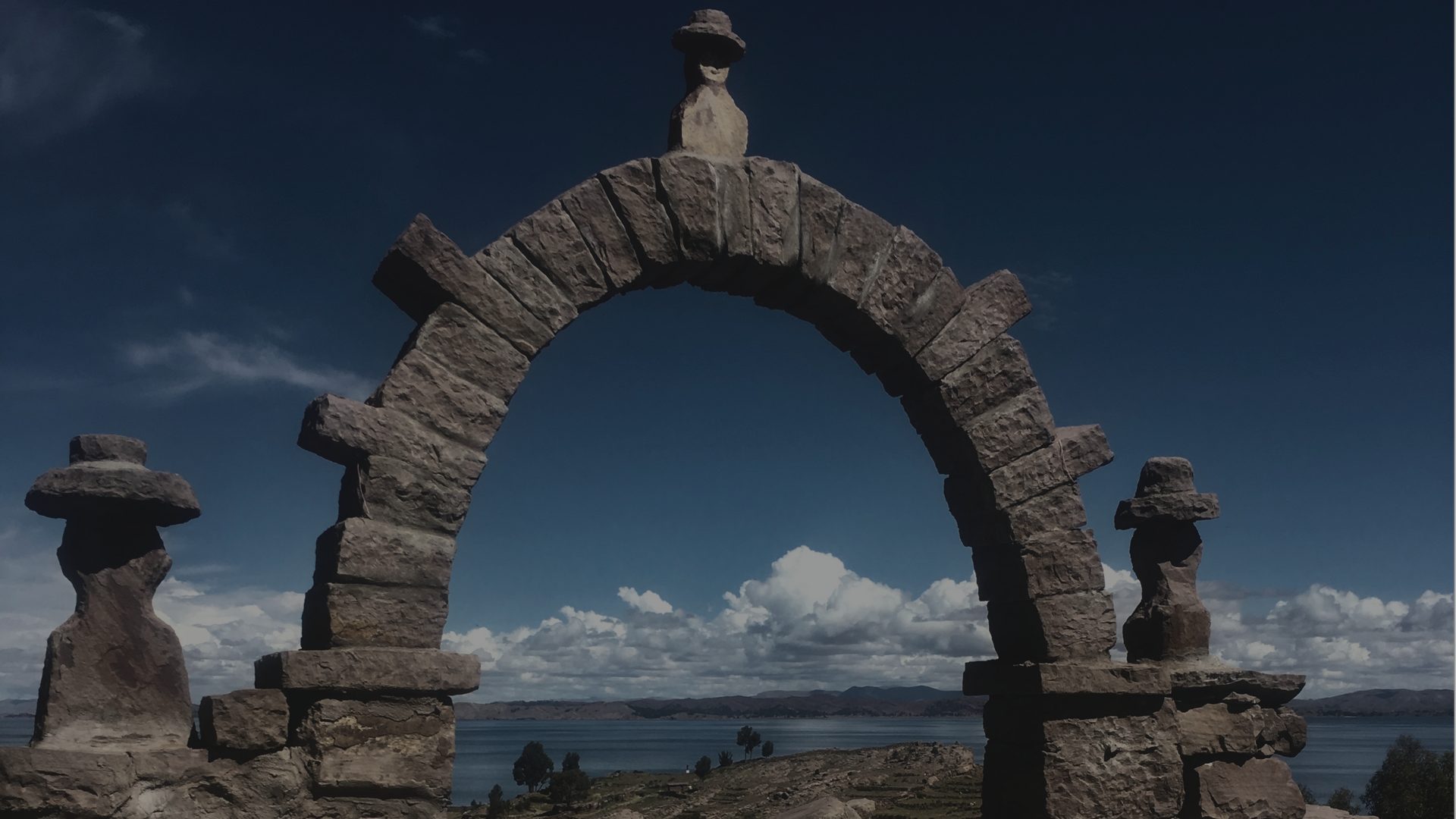 A stone archway frames a distant lake and clouds, flanked by two small stacked-stone cairns.
