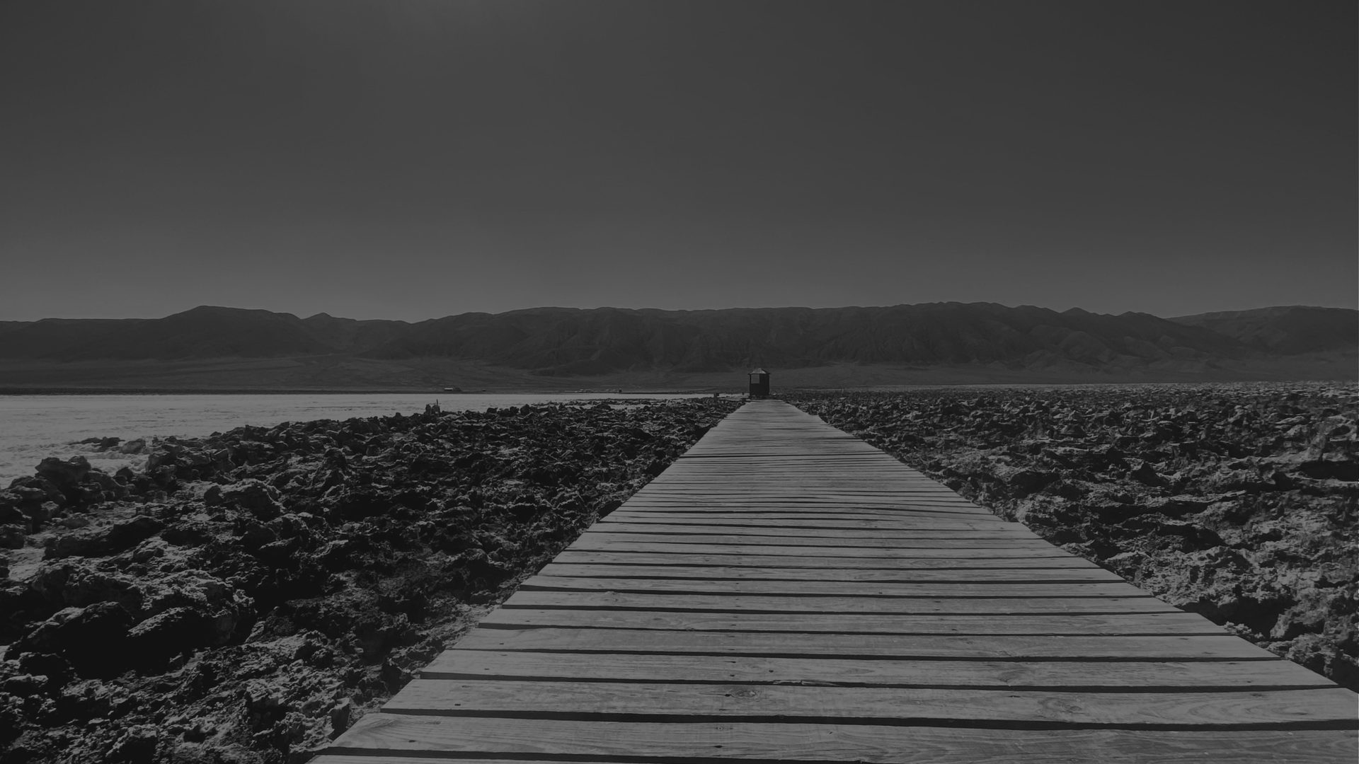 A wooden boardwalk runs straight toward low mountains across a dark, cracked salt flat.
